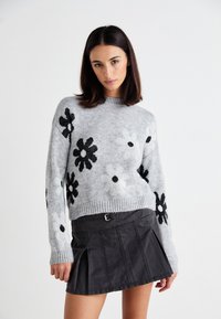Grauer Strickpullover mit schwarzen floralen Mustern, Rundhals-Ausschnitt und geripptem Saum. Kombiniert mit einem schwarzen geplissten Rock, der über einen Gürtel verfügt.