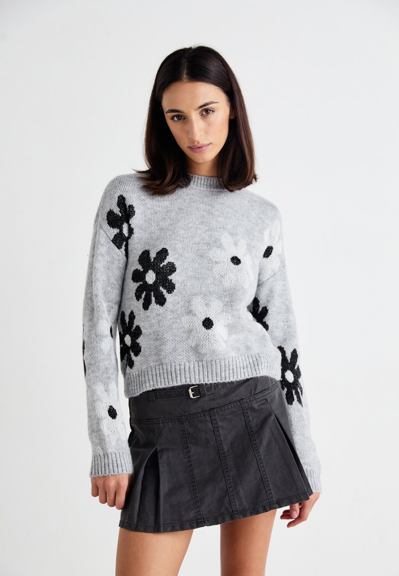 Grauer Strickpullover mit schwarzen floralen Mustern, Rundhals-Ausschnitt und geripptem Saum. Kombiniert mit einem schwarzen geplissten Rock, der über einen Gürtel verfügt.