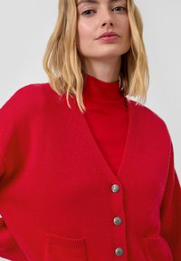 Rote Strickjacke mit V-Ausschnitt, Knopfleiste und zwei vorderen Taschen; verfügt über silberne Knöpfe und gerippte Bündchen. Weiche Textur.