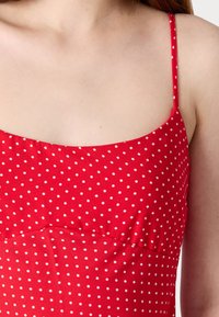Raudona su baltais taškeliais suknelė su plonomis petnešėlėmis, turinti prigludusį liemenį ir lengva raukšlė pasisioje. Lygi medžiagos tekstūra, apvalus iškirpimas.