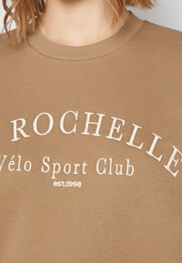 Světle hnědý sweatshirt s kulatým výstřihem a bílým vyšitým textem "ROCHELLE Vélo Sport Club est.1998" na hrudi.