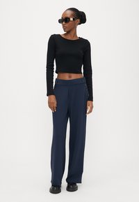 Kvinne iført svart langermet crop-top, marineblå høytlivs vide bukser med knapper, svarte solbriller og svarte sko, stående mot hvit bakgrunn.