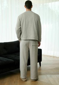 Man in gray sweatpants and sweatshirt standing barefoot on wooden floor near black couch and sheer white curtains.