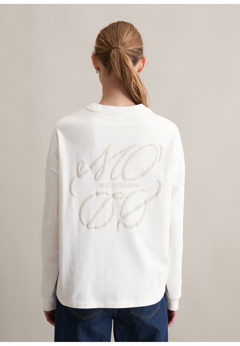 Person mit hellbraunen Haaren im Pferdeschwanz trägt ein weißes Langarmshirt mit eingeprägtem "Marc O'Polo Denim"-Text auf dem Rücken.