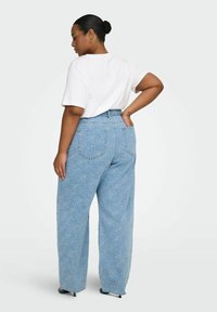 Femme avec cheveux en chignon portant un t-shirt blanc et un jean large taille haute bleu clair à motif de roses, debout, une main sur la hanche.