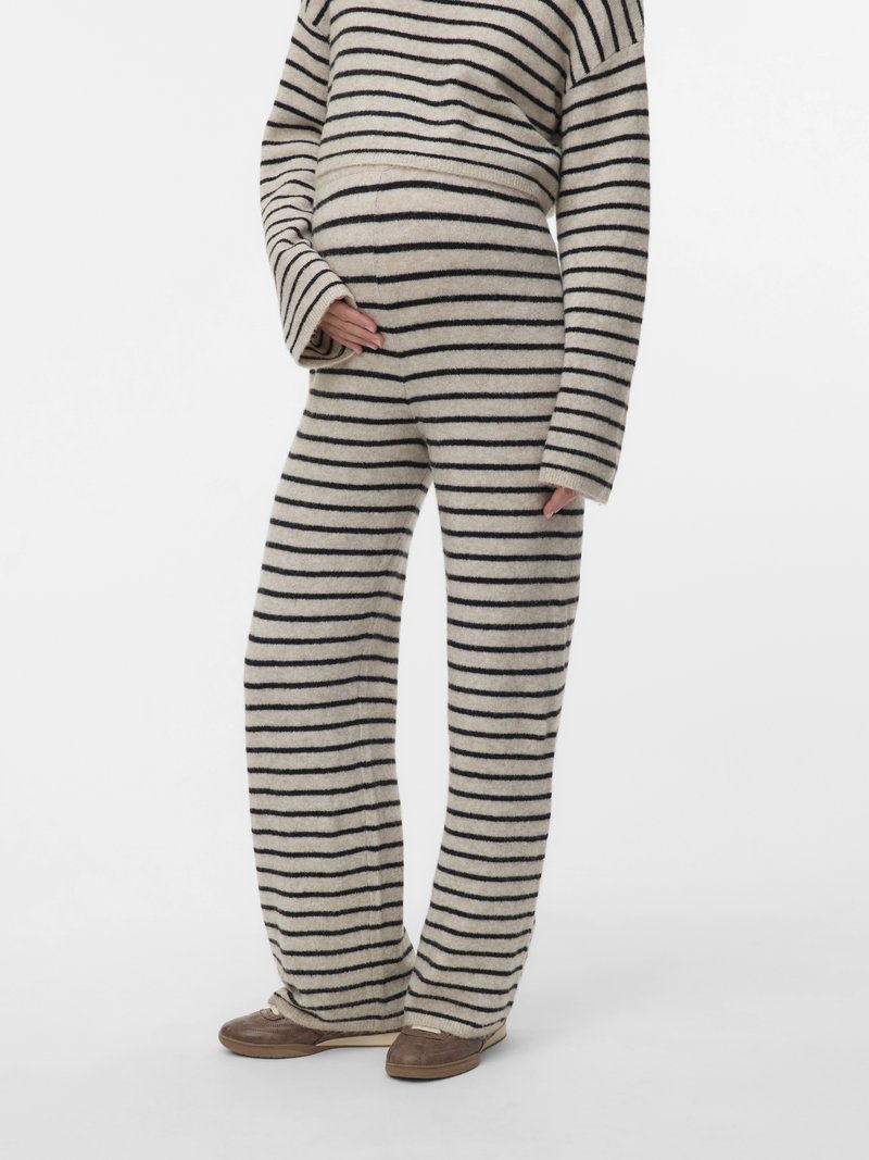 Άτομο με ζεστή πλεκτή ενδυμασία μπεζ και μαύρες ρίγες που κρατά απαλά την έγκυο κοιλιά του, φορά καφέ παπούτσια, στέκεται σε λευκό φόντο.