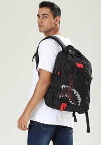 Sac à dos noir avec des accents rouges, présentant un design de requin sur l'avant, plusieurs compartiments et des bretelles rembourrées.