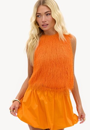 Femme blonde portant un haut orange sans manches duveteux et une jupe orange assortie, tenant les bords de la jupe, fond blanc.