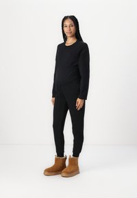 Sudadera negra de maternidad con cintura elástica y pantalones negros a juego. La modelo lleva botas de ante en color beige hasta el tobillo, con suela gruesa y forro de piel.