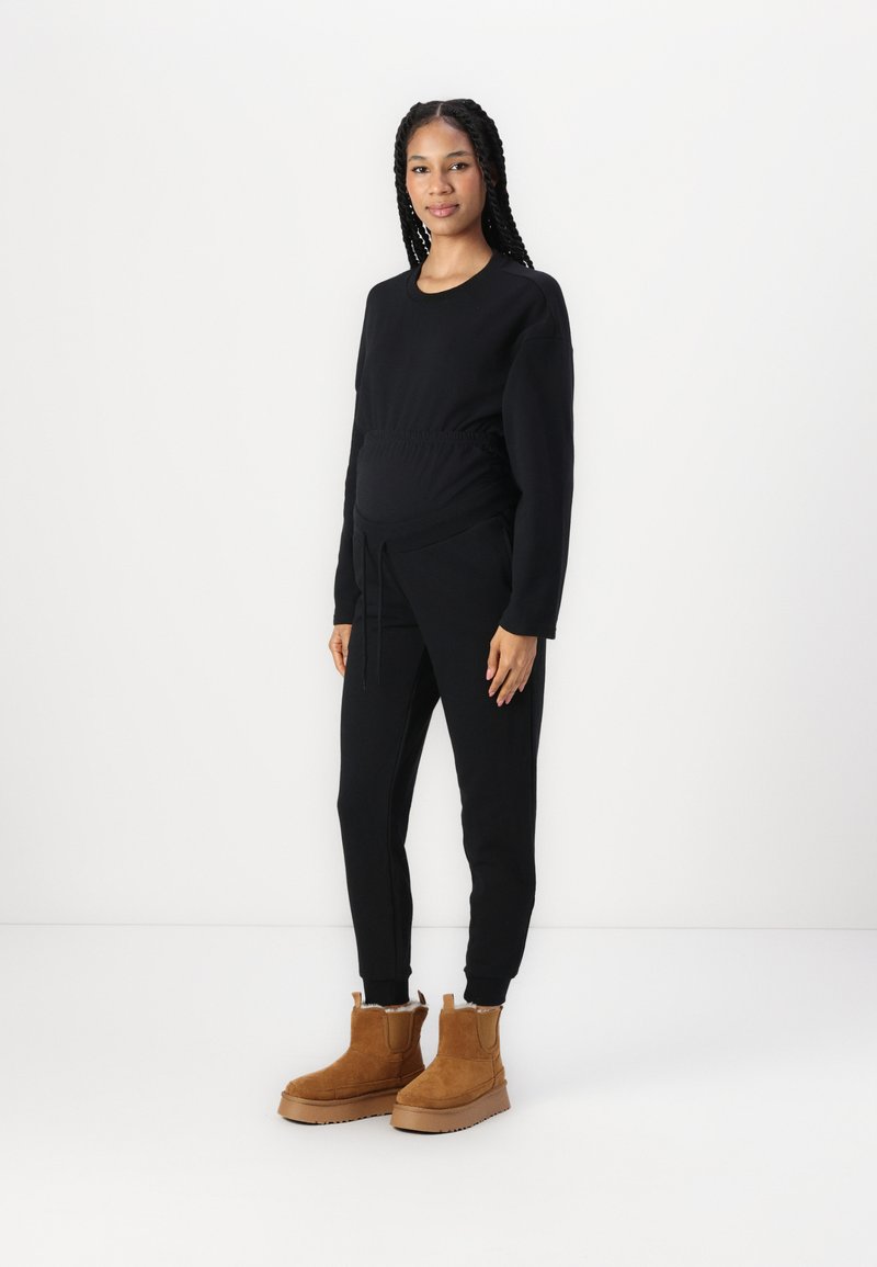 Sudadera negra de maternidad con cintura elástica y pantalones negros a juego. La modelo lleva botas de ante en color beige hasta el tobillo, con suela gruesa y forro de piel.