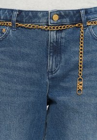 Jean en denim bleu avec une ceinture chaîne dorée passée dans les passants, avec un pendentif doré suspendu près de la poche avant droite.