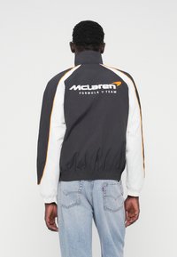 Μαύρο και άσπρο αδιάβροχο μπουφάν με πορτοκαλί λεπτομέρειες, με το λογότυπο "McLaren Formula 1 Team" σε άσπρο στην πλάτη, ελαστικά μανίκια και τελείωμα.