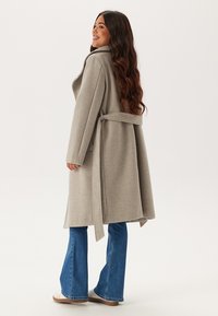 Cappotto di lana grigio chiaro con vita cintrata, maniche lunghe e un design fluido, abbinato a jeans svasati e sneakers.