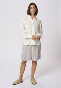 Lichte, lange mouwen witte blouse met een knoopsluiting aan de voorkant, gecombineerd met knielange strepen shorts in beige en wit. Draagt met beige klompen.