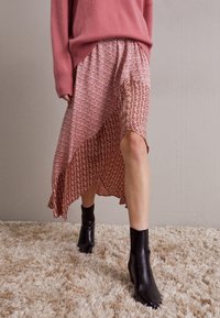 Maxi-Rock aus gemustertem Stoff mit asymmetrischem Saum, kombiniert mit schwarzen Stiefeletten. Das Outfit umfasst einen pinken strukturierten Pullover.