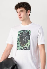 Fehér póló, amelyen egy fekete párduc grafika látható zöld lombozattal, aranylánccal a nyakában, és a "BOSS" logó hangsúlyosan megjelenítve.