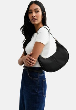Zwarte faux leren schoudertas met een gebogen vorm, ritssluiting en verstelbare schouderband, gedragen tegen een wit t-shirt en blauwe jeans.