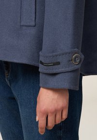 Giacca in misto lana di colore blu scuro, con polsini con dettagli a bottoni e un'etichetta con la scritta "Cucito a Mano". Jeans visibili sotto.