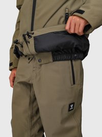 Persona che sistema l'orlo di una giacca verde oliva con un rivestimento interno nero, indossando pantaloni verde oliva abbinati con tasche con zip.