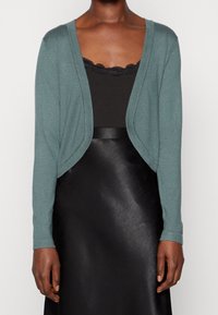 Teal stickad cardigan med öppen frontdesign och långa ärmar, bärs över en svart topp och en svart kjol i imitationsläder.