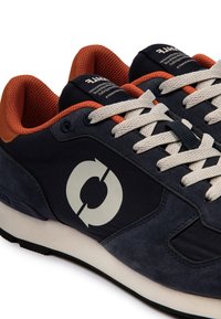 Baskets bleu foncé avec des lacets blancs, une doublure intérieure orange, et un logo blanc circulaire sur le côté, vues en trois quarts.