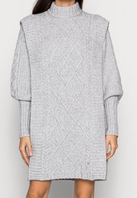 Grå stickad klänning med hög krage, puffärmar, kabelstickat mönster, ribbade detaljer och en liten logotyp nära fållen.