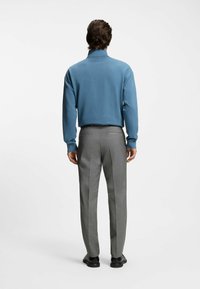 Lichtblauwe trui voor heren met een hoge kraag, gecombineerd met grijze, op maat gemaakte broek. De stof oogt glad; de schoenen zijn zwart en gepolijst.