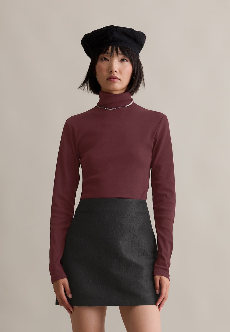 Bordeaux langemouw turtleneck shirt gecombineerd met een zwarte textuur mini-jurk en een zwarte baret, geaccentueerd met een zilveren ketting.