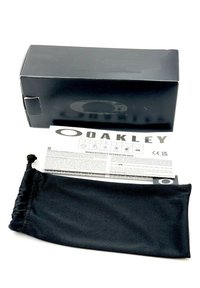 Scatola degli occhiali Oakley nera con foglio informativo accanto a una custodia in microfibra nera, dotata di chiusura a cordoncino e accentazione del logo.