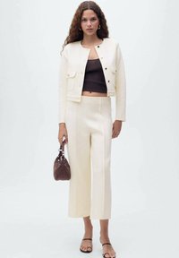 Cremfarbener Blazer mit goldenen Knöpfen und Fronttaschen, kombiniert mit cremfarbenen weitem Hosen. Die Schuhe sind schwarze Riemchensandalen; sie hält eine braune Handtasche.