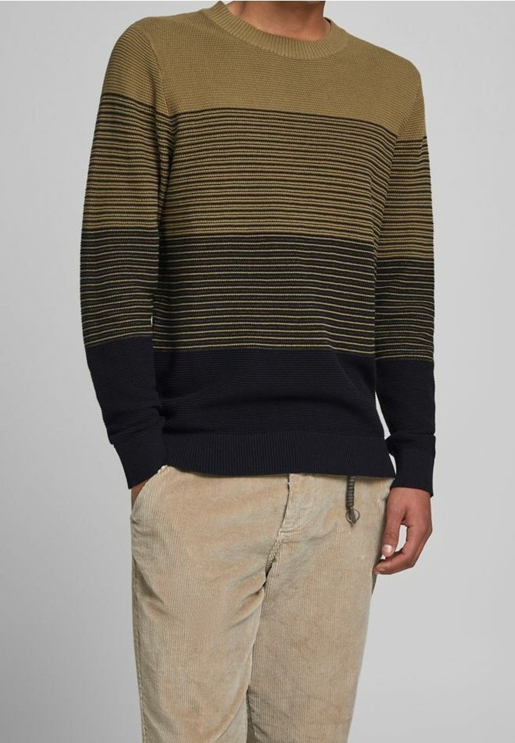 Homme portant un pull en tricot rayé marron et noir avec un pantalon en velours côtelé beige, debout avec une main partiellement dans la poche.