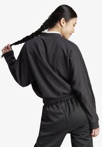 Črna majica s kapuco, ohlapnega kroja in spušenimi rameni, v kombinaciji s črnimi trenirkami, ki imajo elastiko v pasu in gladko teksturo.