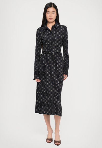 Abito nero a maniche lunghe con motivo a spirale bianco, colletto, vita con cintura, orlo svasato e tessuto testurizzato. Indossato con scarpe a punta scoperta marroni.