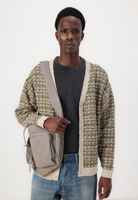 Strikket cardigan i olivengrøn med cremfarvet mønster, t-shirt med rund hals, lyseblå jeans og grå crossover taske med lynlås.