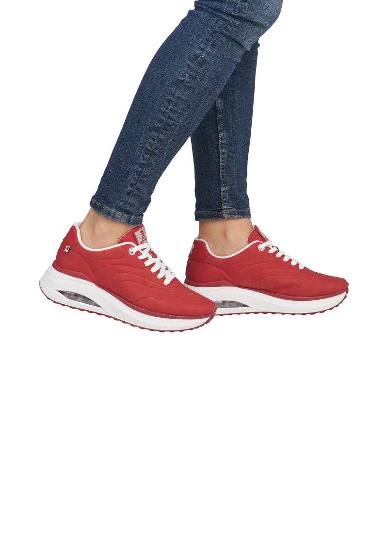 Red athletic shoes with a mesh upper, white laces, and cushioned sole featuring a visible air unit. Worn with dark blue fitted jeans.