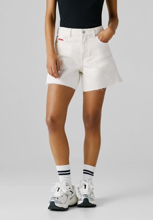 Isik, kes kannab valgeid teksapükse roosteta servaga, musta topi, valgeid sokke mustade triipudega ning valgeid spordijalatseid, seisab ühevärvilise tausta ees.