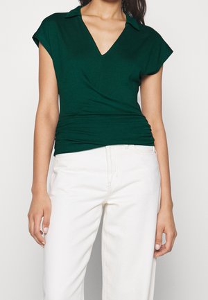 Vrouw die een donkergroen kort mouw shirt met V-hals en kraag draagt, gecombineerd met een hoge taille witte broek, staande tegen een effen achtergrond.