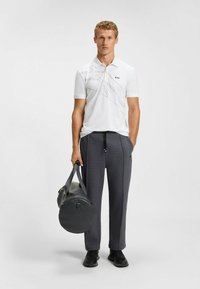 Weißes Polohemd mit einem Linienmuster und dem 'BOSS'-Logo, kombiniert mit dunkelgrauen, schmal zulaufenden Hosen, während eine schwarze runde Sporttasche gehalten wird.