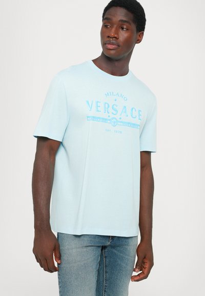 Light blue cotton t-shirt with short sleeves, featuring a central printed logo design in darker blue. Straight hem and relaxed fit.