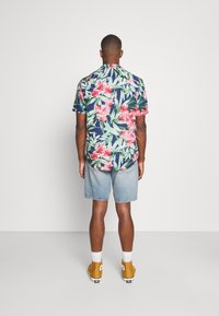 Kurzärmeliges Blumenhemd mit blauem, rosa und grünem Muster; kombiniert mit Jeansshorts und gelben Turnschuhen. Schlichter Hintergrund.