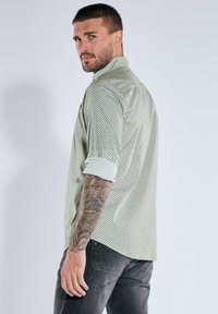 Mann mit kurzem Haar und Tätowierungen am linken Arm trägt ein hochgerolltes gemustertes Hemd und dunkle Jeans und schaut über die Schulter zurück.