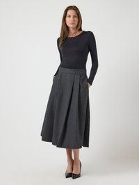 Mujer con cabello largo vestida con una blusa negra de manga larga, falda midi plisada gris oscuro, tacones negros, de pie frente a un fondo claro y liso.