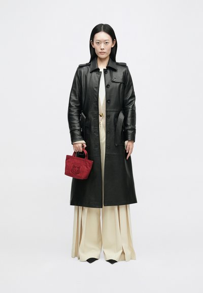 Casaco longo de couro preto com botões à frente, bolsos laterais e cintura com cinto. Combinado com calças largas bejes e uma mini bolsa vermelha com bordados.