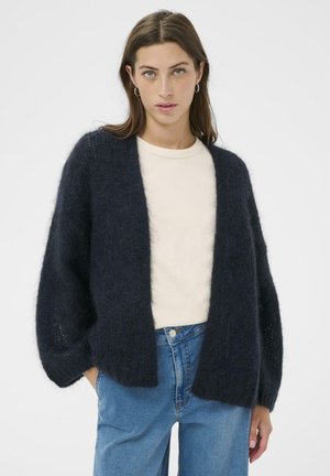 Dark navy knitted cardigan with wide, textured sleeves and an open front, worn over a cream shirt and light blue jeans. Soft, plush material.