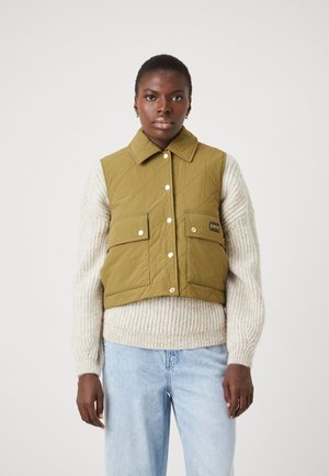 Gepolsterte olivgrüne Weste mit Kragen, Frontdruckknöpfen, zwei Brusttaschen und einem schwarzen Etikett; getragen über einem cremefarbenen gerippten Pullover.