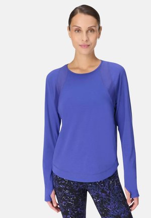 Vrouw draagt een blauwe sporttop met lange mouwen en zwart-paarse legging met patroon, staand tegen een lichtgrijze achtergrond.