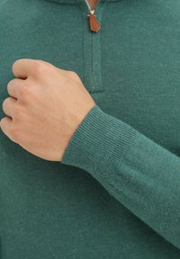 Pull à col zippé vert en matière douce, doté de poignets côtelés et d'une tirette en cuir, mettant en valeur une surface lisse et texturée.