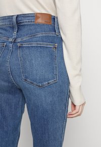 Jeans de mezclilla azul con cintura alta. Presentan un parche de cuero marrón, dos bolsillos traseros y costuras en contraste a lo largo de las costuras.