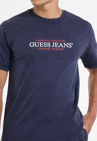 Granatowa bawełniana koszulka z krótkimi rękawami. Ozdobiona grafiką na przodzie z białym i czerwonym tekstem: "AMERICAN TRADITION GUESS JEANS ORIGINAL DESIGNS."