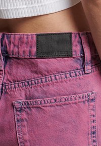 Rosa denimjeans har framträdande sömmar, en svart läderetikett med "SUPERDRY"-logotyp samt en klassisk femficksdesign.